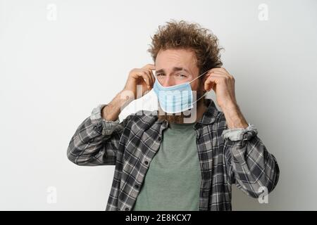 Déguisement sous la contrainte met sur son visage un masque médical de protection pour protéger le coronavirus, avec les cheveux bouclés, jeune homme portant une chemise à carreaux et olive Banque D'Images