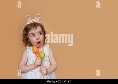 Portrait positif de petite fille avec des oreilles de lapin sur la tête, regarde dans la surprise, tenant un oeuf vert orange dans ses mains. Arrière-groupe orange clair isolé Banque D'Images