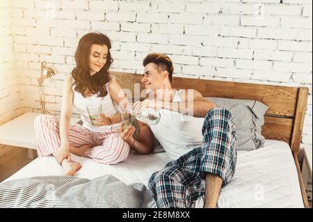 Joyeux couple de youmg en pyjama avec des verres d'eau sur lit à la maison Banque D'Images