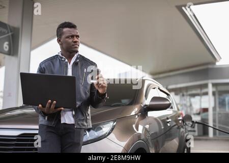 Homme d'affaires noir à la station-service, debout près de la voiture avec un café à emporter et un ordinateur portable et regardant loin, tout en faisant le plein de son auto. Station-service Banque D'Images