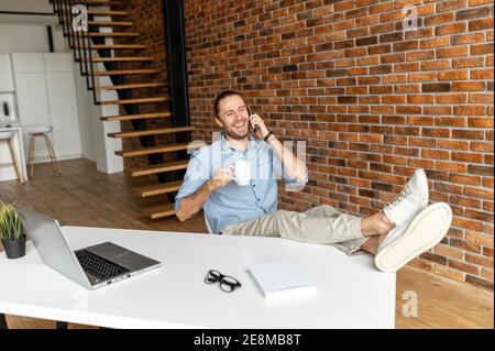 Jeune entrepreneur détendu du millénaire assis dans le bureau à domicile avec ses jambes sur le bureau, a terminé le projet et prendre une pause café, tenant Banque D'Images