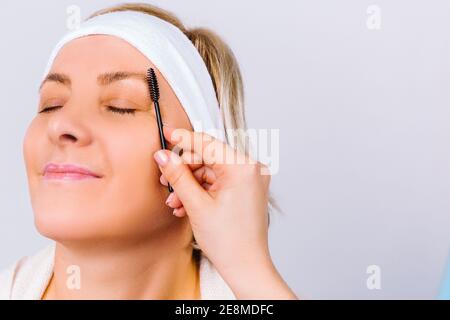 Femme âgée avec une peau phosphorescente et un maquillage doux et la main du maître fait une lamination des sourcils sur un fond blanc. Côté vide pour la beauté Banque D'Images