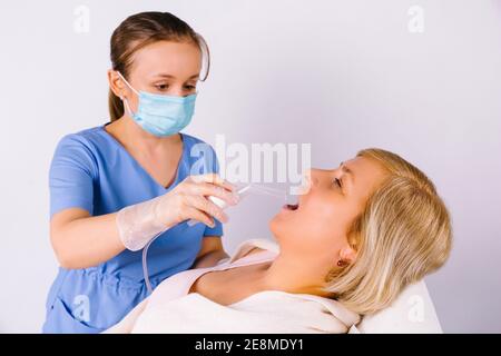 Un jeune médecin portant un masque médical et des gants en latex traite la gorge d'une femme plus âgée avec un nébuliseur sur fond blanc. Lutte contre le rhume des virus. Banque D'Images