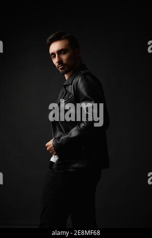 Jeune homme beau avec visage non rasé dans une veste en cuir noir posant dans le fond sombre, isoler. Style barbu dans une veste en cuir Banque D'Images