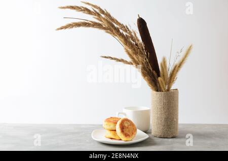 Quelques crêpes sur une assiette blanche et une tasse vide sur une table grise texturée contre un mur blanc avec un bouquet d'automne d'herbes séchées et de roseaux. Sélectionnez Banque D'Images