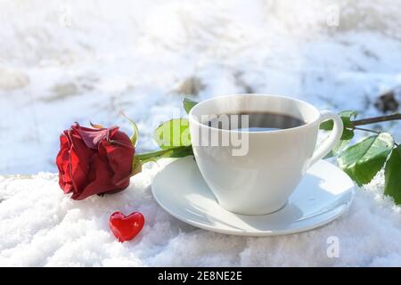 Café chaud dans une tasse blanche servi à l'extérieur dans la neige, décoré avec une rose rouge et un petit coeur de verre lors d'une belle journée d'hiver, concept d'amour pour Banque D'Images