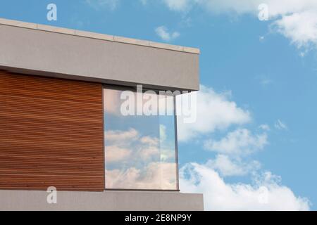 Détail d'une maison moderne en cube avec panneaux en bois et ciel bleu avec des nuages blancs reflétant dans les grandes fenêtres. Banque D'Images