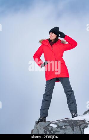 Jeune femme debout sur le sommet de la montagne sur fond de ciel avec des nuages spectaculaires. Femme souriante vêtue d'une veste coupe-vent rouge, d'un pantalon de sport Banque D'Images