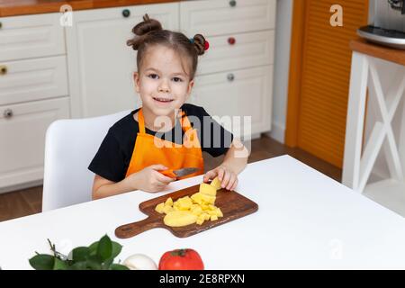 petite fille assise à la table à l'arrière-plan de la cuisine tout en coupant légumes Banque D'Images