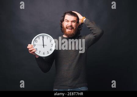 Un jeune homme barbu regarde stressé à la caméra tenant une horloge murale . Banque D'Images