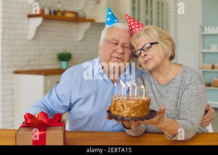Joyeux couple d'âge mûr en chapeaux de cône soufflant des bougies sur le gâteau célébrant l'anniversaire à la maison. Banque D'Images