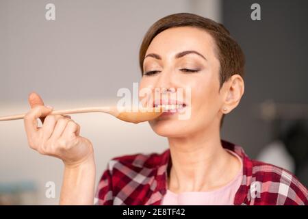 Gros plan d'une femme essayant un plat cuit à l'aide d'une longue cuillère en bois. Femme avec un cheveux court de cuisine un dîner pour la famille debout dans la cuisine moderne Banque D'Images