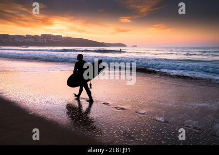 Un surfeur portant sa planche de surf et marchant le long du rivage sur la plage de Fistral silhoueté par le coucher du soleil à Newquay, dans les Cornouailles. Banque D'Images