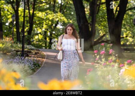 une jeune femme vêtue d'un haut clair et d'une jupe variable marche à travers le parc de la ville, des fleurs défoquées en premier plan Banque D'Images