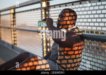 Portrait du jeune homme afro-américain en vêtements de sport qui boit de l'eau après l'exercice. Il fait de l’exercice pour réduire son poids. Banque D'Images