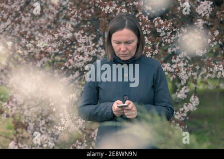 La messagerie texte de la femme sur le téléphone portable sous la cerise fleurie arbre au printemps Banque D'Images