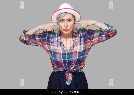 Je ne veux pas vous entendre. Portrait de femme mature moderne et élégant dans un style décontracté avec white hat standing, mettre les doigts dans les oreilles et à la recherche. Banque D'Images