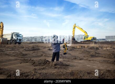 garçon de 4 ans dans des vêtements chauds à un le chantier de construction avec son dos dans le cadre tient un pelle et regarde les gros équipements de construction Banque D'Images