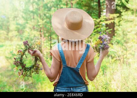 Femme dans un chapeau de paille et une combinaison en denim tient une couronne de fleurs de forêt. Vue arrière. Arrière-plan de la forêt Banque D'Images