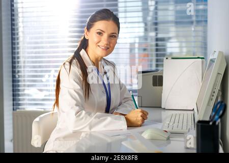 femme médecin utilisant un ordinateur tout en étant assis au travail, posant à la caméra, elle est un lieu de travail de médecin. santé, assurance et medic Banque D'Images