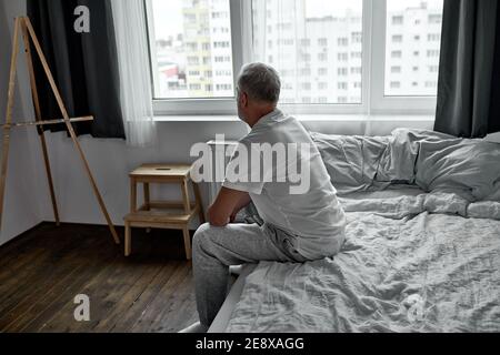 vue latérale sur l'homme contrarié assis seul sur le lit à la maison dans la pièce sombre, le matin seul. Banque D'Images