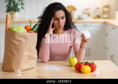 Dépenses d'épicerie. Femme préoccupée assise dans la cuisine, regardant le projet de loi d'achat de nourriture Banque D'Images