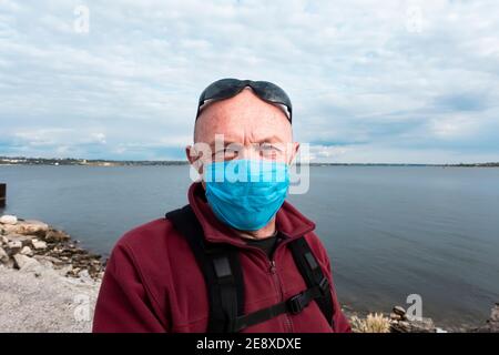 un homme chauve dans un chandail rouge avec un masque de protection médicale et des lunettes de soleil se tient sur la rive de la rivière. Un homme âgé dans une période d'auto-isolement et q Banque D'Images