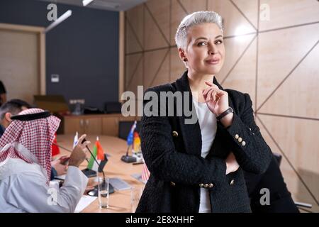 femme d'affaires agréable avec des cheveux courts en tenue formelle posant dans salle de réunion lors d'une réunion avec un groupe international de politiciens bureau Banque D'Images