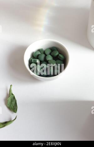 Pilules de spiruline verte dans un bol sur fond blanc, composition minimaliste avec supplément alimentaire végétalien organique végétal et arc-en-ciel de lumière, gros plan, il Banque D'Images