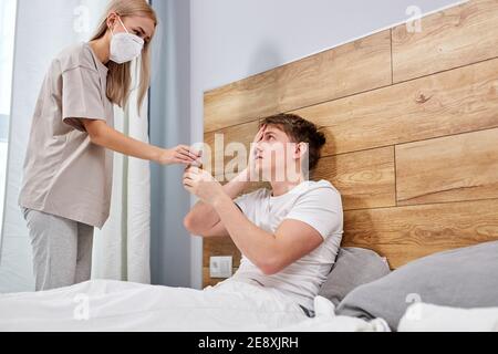 homme ayant froid, allongé sur le lit à la maison se sentant mal et femme dans le masque médical prend soin de donner des médicaments, la rhinite et l'allergie saisonnière, la grippe Banque D'Images