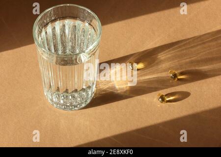 Un verre d'eau avec des capsules d'huile de poisson sur fond marron terre avec lumière et ombres, gros plan, espace de copie, supplément sain et conce de traitement Banque D'Images