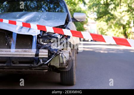 Voiture détruite dans un accident de la circulation sur la route de la ville avec ruban rouge d'avertissement de police. Pare-feu avant automatique cassé, capot bosselé sans pare-chocs Banque D'Images