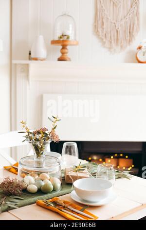 Table en bois blanc avec assiette et bol blancs, œufs de Pâques pastel, fleurs de jardin fraîches et cadeau brun enveloppé de papier, décor de salon Banque D'Images