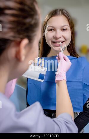 Portrait en gros plan d'une belle jeune femme assise sur une chaise dentaire tandis que les mains de stomatologiste dans des gants stériles tenant des échantillons de dents. Banque D'Images