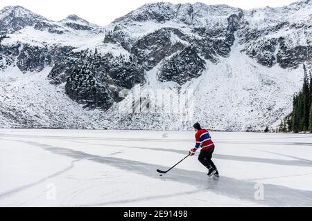 Vue arrière d'un homme anonyme en uniforme jouant au hockey sur glace sur un lac gelé près de la pente enneigée de la montagne en hiver dans la campagne de la Colombie-Britannique, CAN Banque D'Images