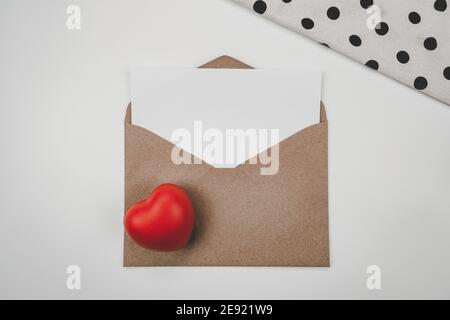 Le papier blanc vierge est placé sur une enveloppe de papier marron ouverte avec un cœur rouge et un chiffon blanc sur fond blanc. Concept de la Saint-Valentin Banque D'Images