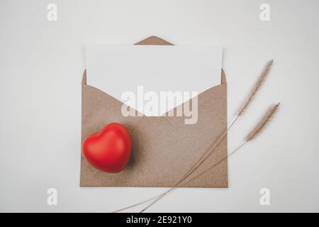 Le papier blanc vierge est placé sur une enveloppe de papier brun ouverte avec un cœur rouge et une fleur sèche de queue d'bœuf sur fond blanc. Concept de la Saint-Valentin. Banque D'Images