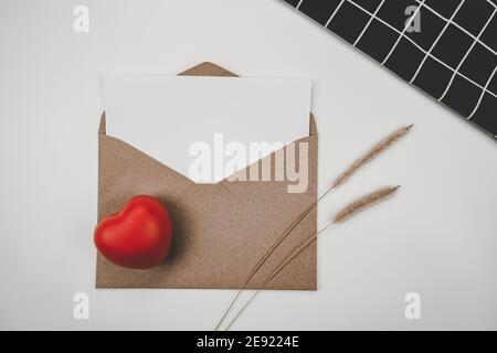 Le papier blanc vierge est placé sur une enveloppe de papier marron ouverte avec un cœur rouge, une fleur sèche et une toile noire sur fond blanc. Saint Valentin Banque D'Images