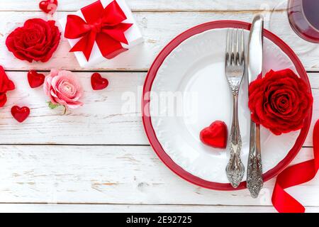 Table romantique de la Saint-Valentin avec une assiette, roses décoratives en papier rouge, cadeau emballé avec un joli noeud et de l'argenterie vintage fantaisie. Banque D'Images