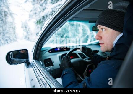 Homme dans des vêtements chauds d'hiver assis dans la voiture. Route de campagne enneigée, voiture couverte de glace, belle forêt sous la neige. Banque D'Images