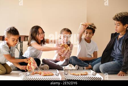 Cinq enfants multiethniques jolis petits garçons et filles assis à l'intérieur manger pizza cuisine italienne fast food. La génération alpha partage les tranches, passez du temps à Banque D'Images