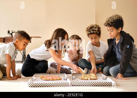 Cinq adorables enfants divers petits garçons et filles s'assoient à l'intérieur en mangeant de la pizza italienne fast food cuisine. La génération alpha partage les tranches, passez du temps à faire des toges Banque D'Images