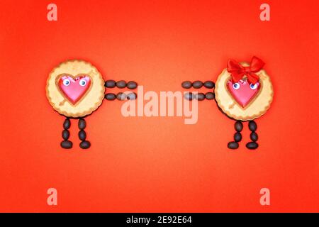 Deux personnages faits de biscuits ronds décorés de coeurs en gelée rouge et de grains de café essayant de se joindre. Couche plate créative isolée sur une base rouge Banque D'Images