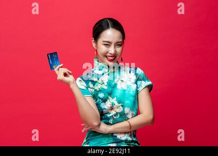 Souriante femme asiatique portant une robe orientale qipao montrant du crédit Carte en main sur fond rouge studio pour nouveau chinois année dépenses sans espèces con Banque D'Images