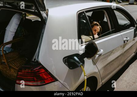 Voiture électrique se charger pendant que la fille regarde par la fenêtre à station-service le jour ensoleillé Banque D'Images