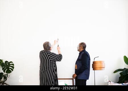 Femme senior martelant l'ongle sur le mur pour fixer la peinture debout par homme âgé dans le salon Banque D'Images