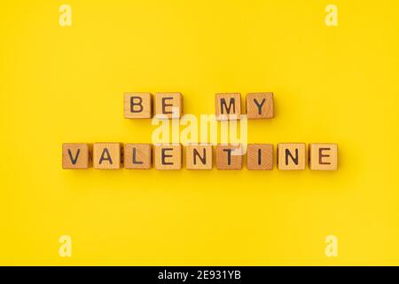 Cubes en bois avec l'expression « soyez ma Saint-Valentin » sur un fond jaune coloré vide. Les mots d'amour sont faits de lettres de bois pour la Saint-Valentin et pour votre petite amie, petit ami, femme, mari saluant carte postale romantique. Flat lay, vue de dessus Banque D'Images