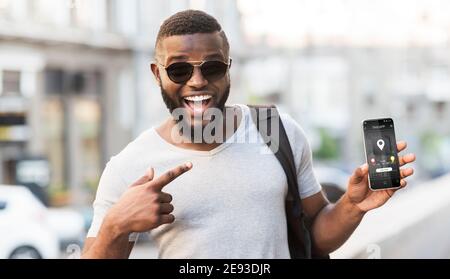 Un gars noir enthousiaste qui pointe vers l'application de taxi mobile sur son téléphone en attendant un taxi sur la rue de la ville, panorama Banque D'Images