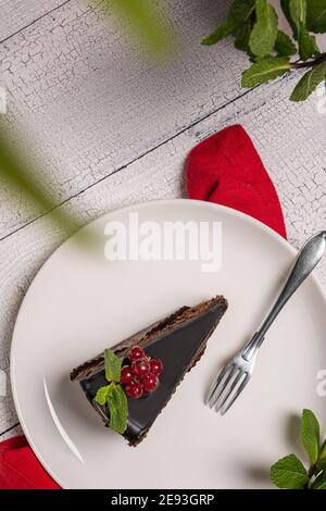 Morceau de gâteau au chocolat appelé Mozart décoré de baies vitrées de cassis rouge et feuille de menthe sur plaque blanche avec fourchette, nappe rouge et sur blanc WO Banque D'Images