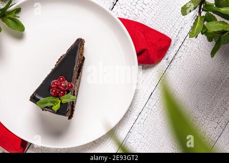 Tranche de gâteau au chocolat appelé Mozart décorée de baies vitrées de cassis rouge et feuille de menthe sur plaque blanche, nappe rouge et bois blanc Banque D'Images